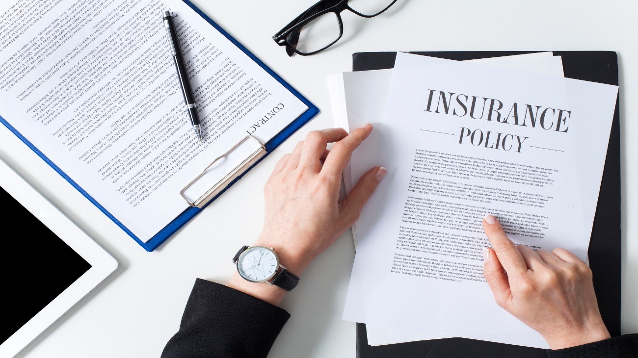 A person reviewing an insurance policy document among other business items like a tablet, glasses, and a clipboard on a white desk.
