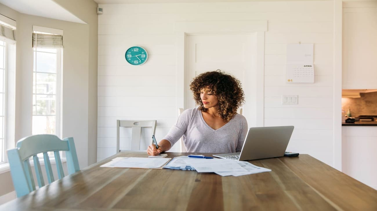 A woman with curly hair sits at a wooden table, working on documents with a laptop beside her in a bright kitchen setting.