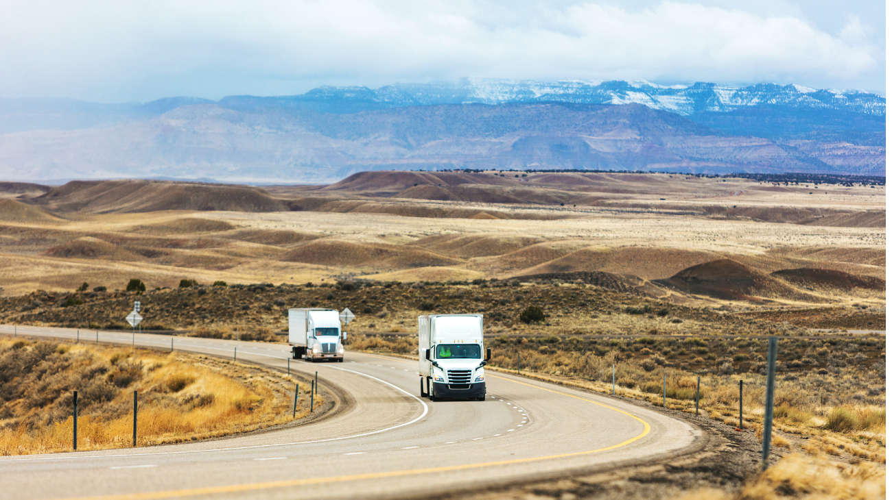 Two trucks driving on a curvy road through a desert landscape with distant snow-capped mountains under a cloudy sky.