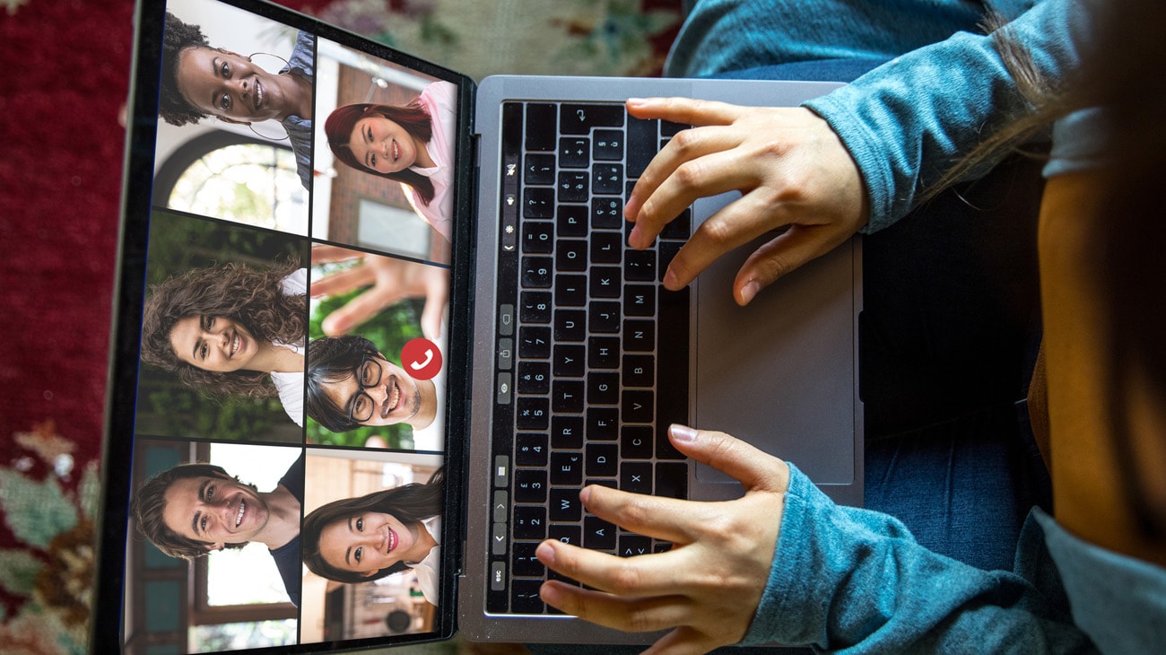 Woman sitting on the floor with a laptop on her lap, the faces of 6 people visible on her laptop on a video call while she manages her team remotely.