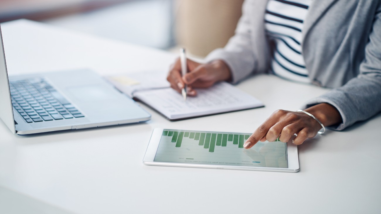 Close-up of a person’s hands analyzing financial data on a tablet, with a notebook and laptop visible on the desk.