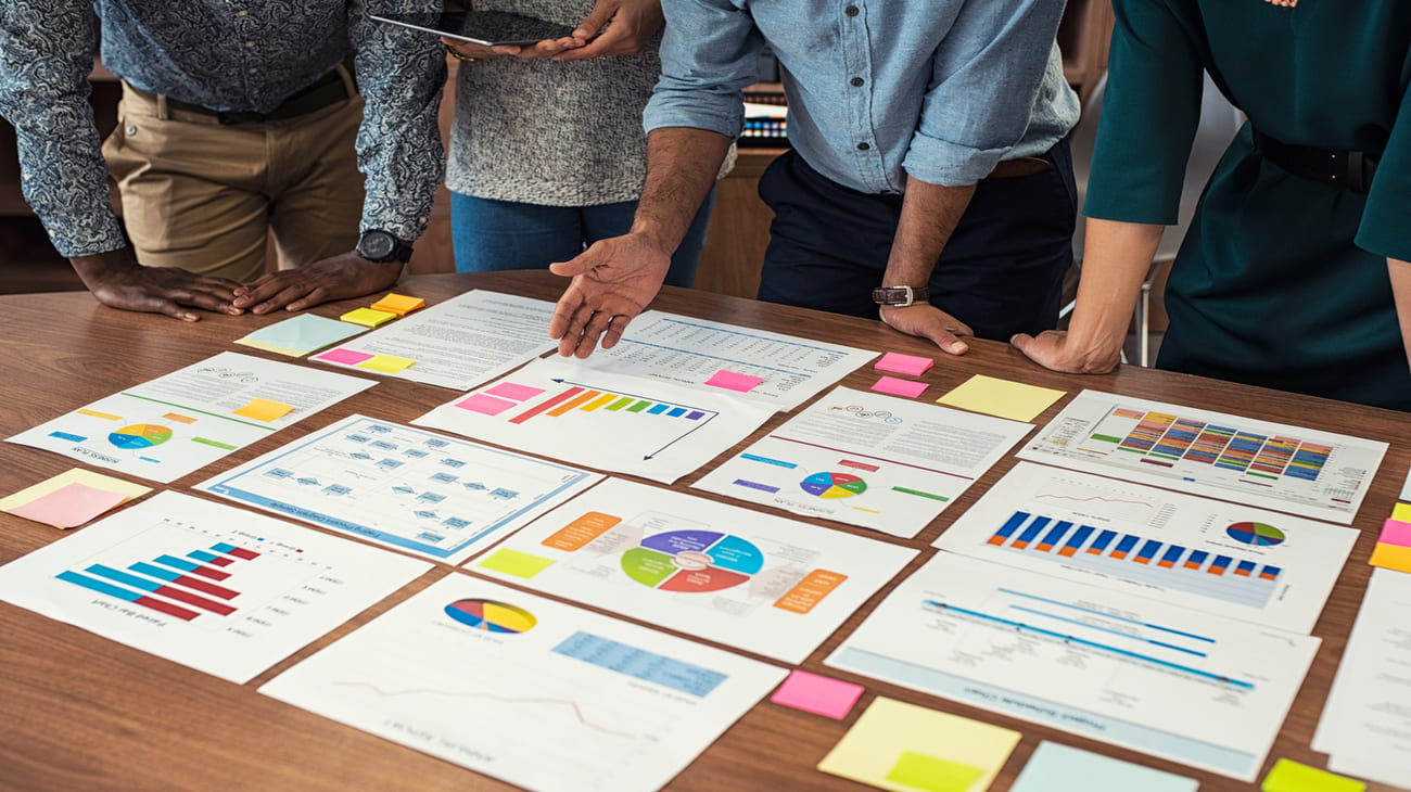 Four professionals standing around a table reviewing various charts, graphs, and documents spread out in front of them.