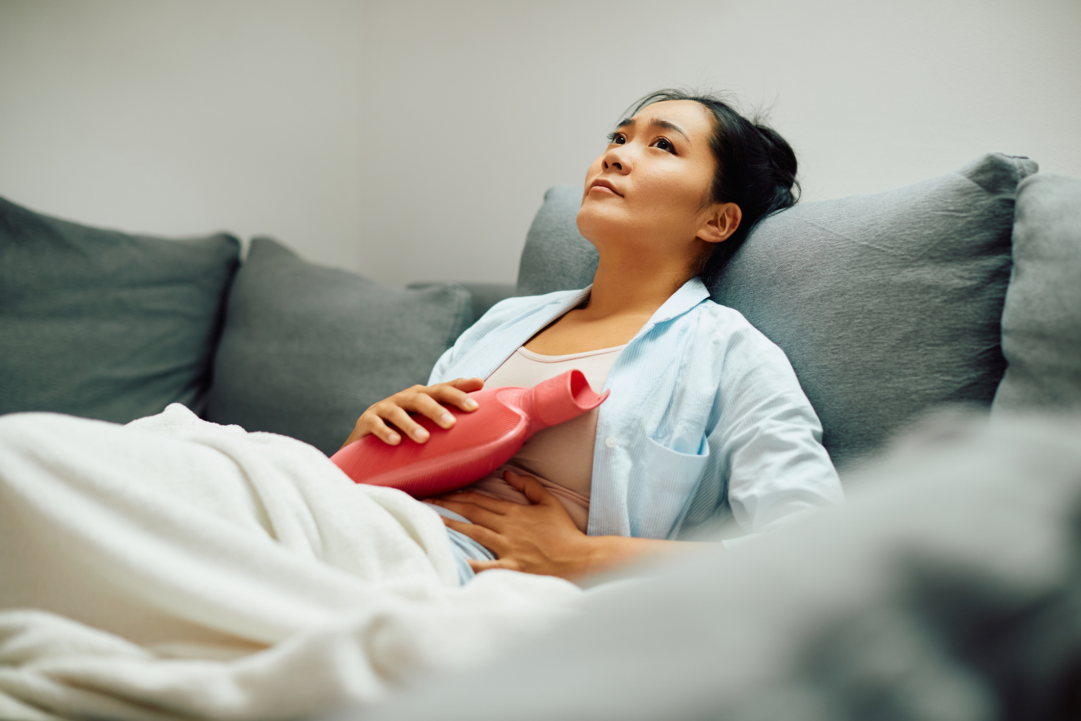 young asian female using a hot watter bottle the alleviate menstrual cramps pain while seated on the sofa