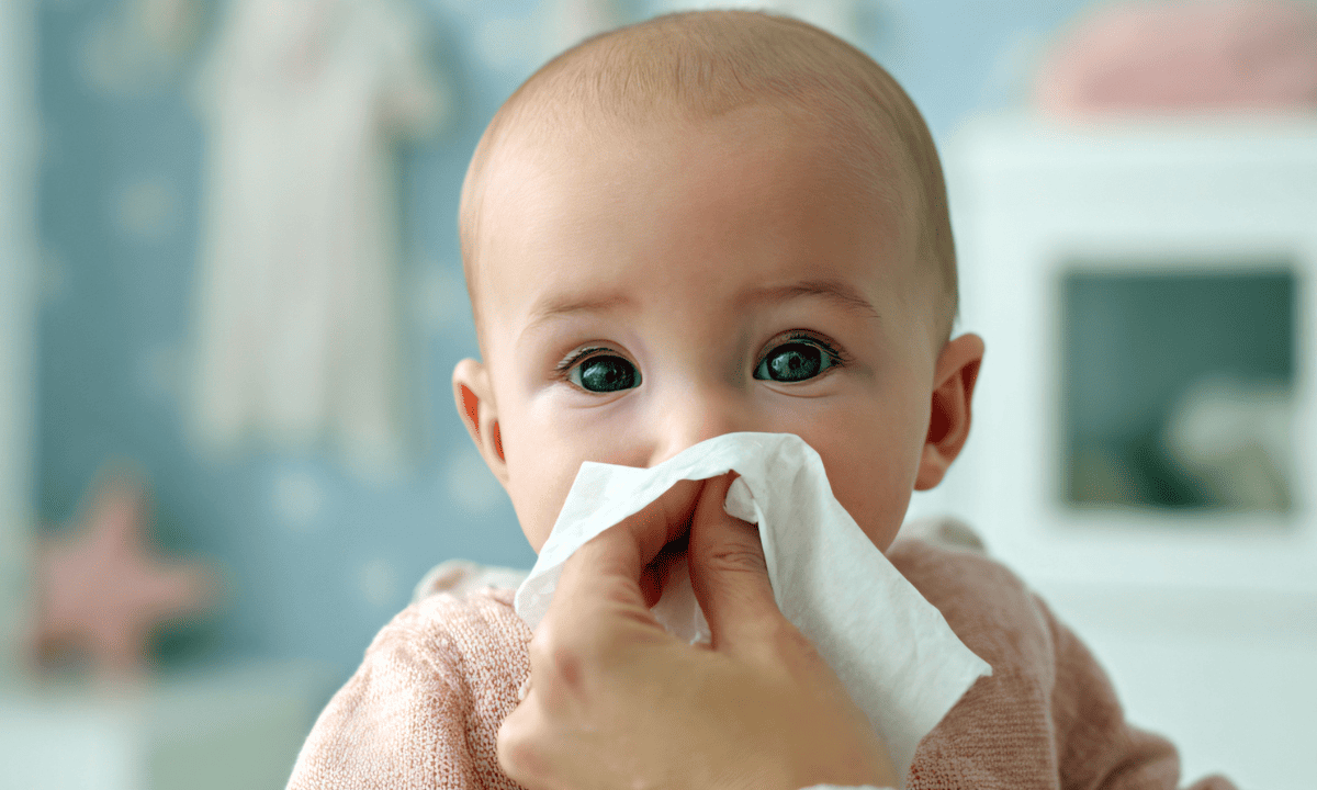 A close-up of an adult wiping a baby's nose with a white tissue.