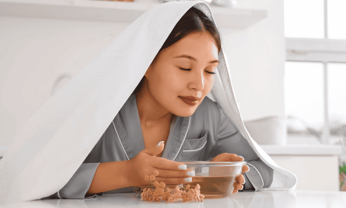 A woman leaning over a bowl of hot water with a towel over her head to inhale steam.