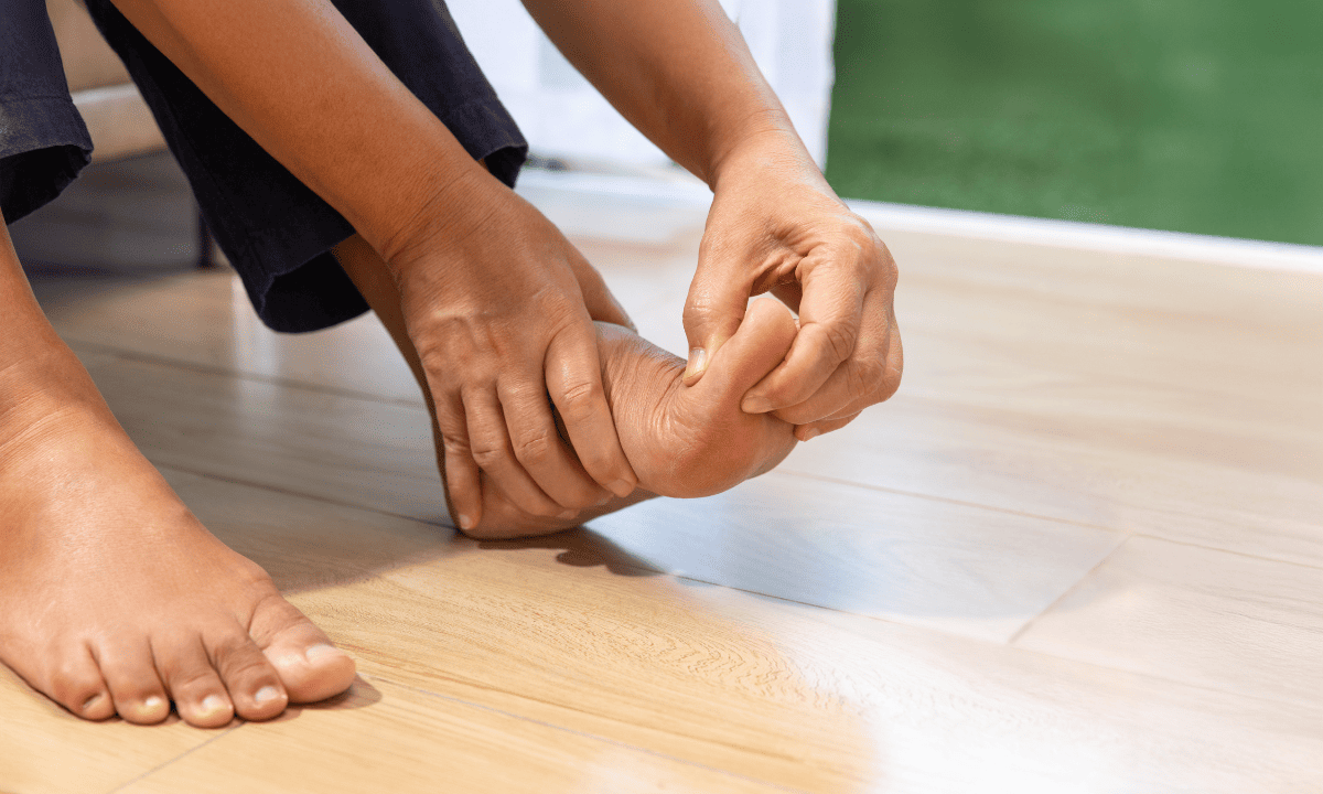 A photo of a person sitting down and holding their foot, specifically touching the base of the big toe, illustrating joint pain or a gout flare-up.