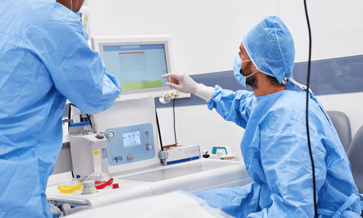 Two medical professionals wearing blue surgical scrubs, caps, and masks working in an operating room, with one pointing at a medical monitor displaying patient data.