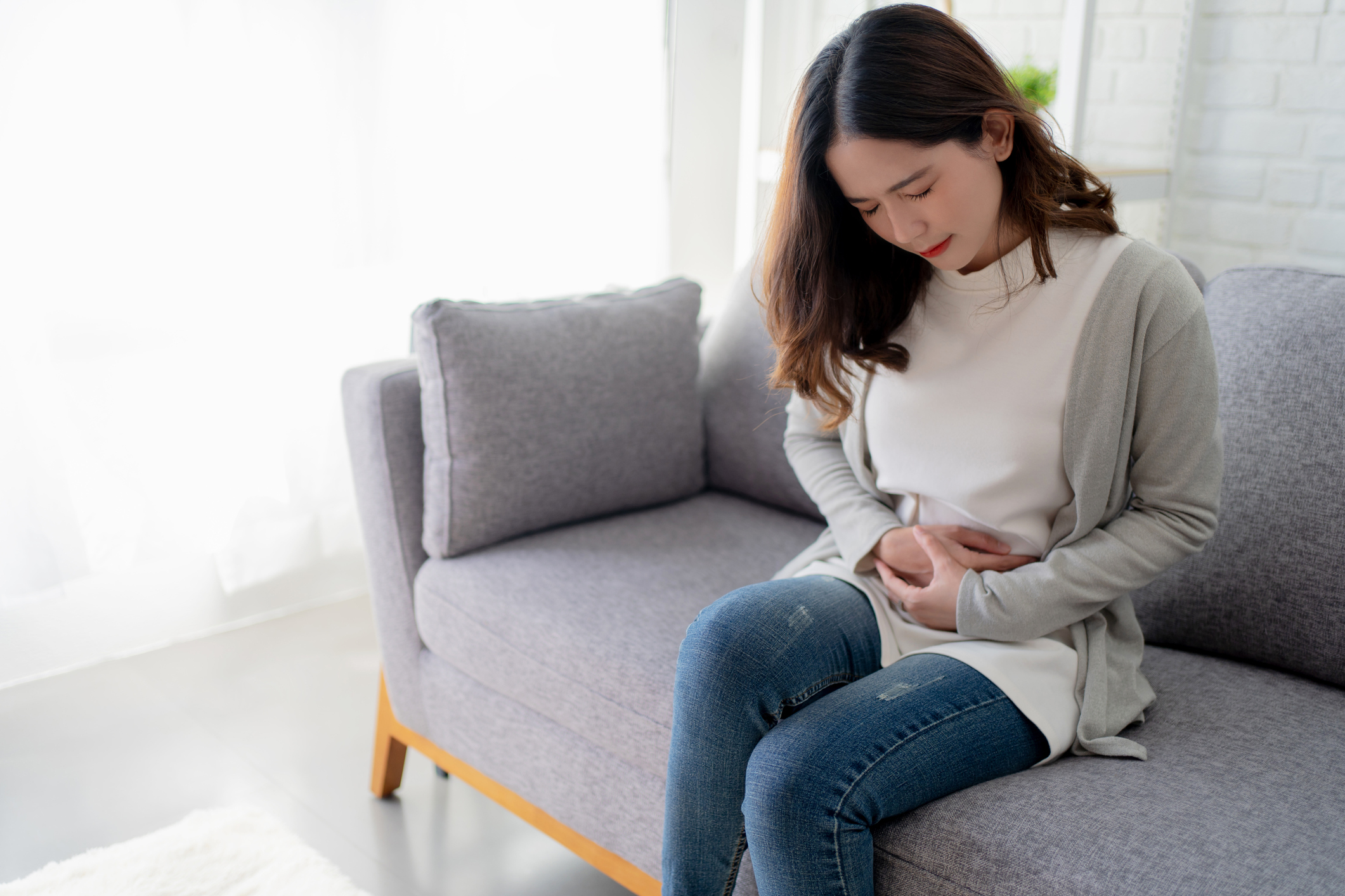 young asian woman sitting on the sofa clutching her abdomen suffering from menstrual cramps and pain pcos