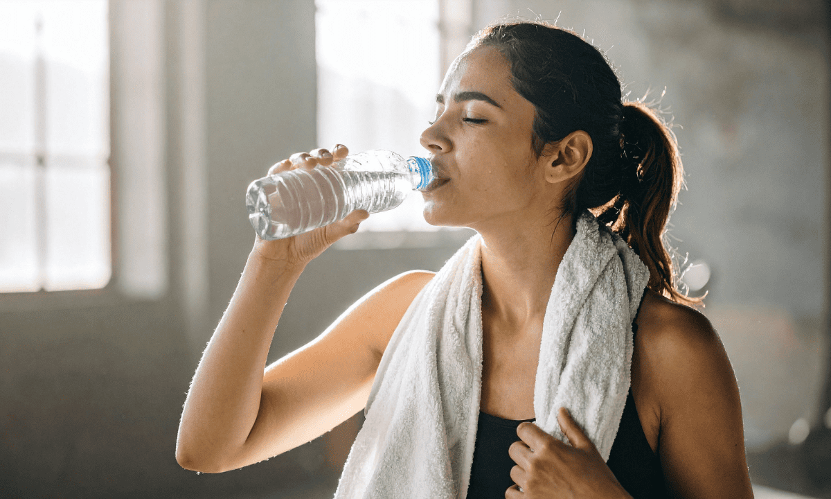 Woman in athletic wear drinking from a plastic water bottle with a towel around her neck.
