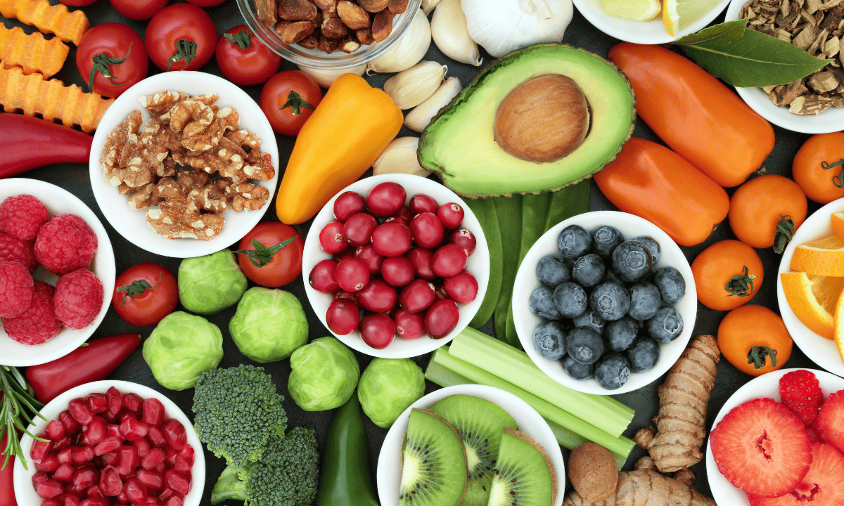 A flat-lay arrangement of fresh, colorful produce including berries, broccoli, peppers, avocado, and walnuts in small white bowls.
