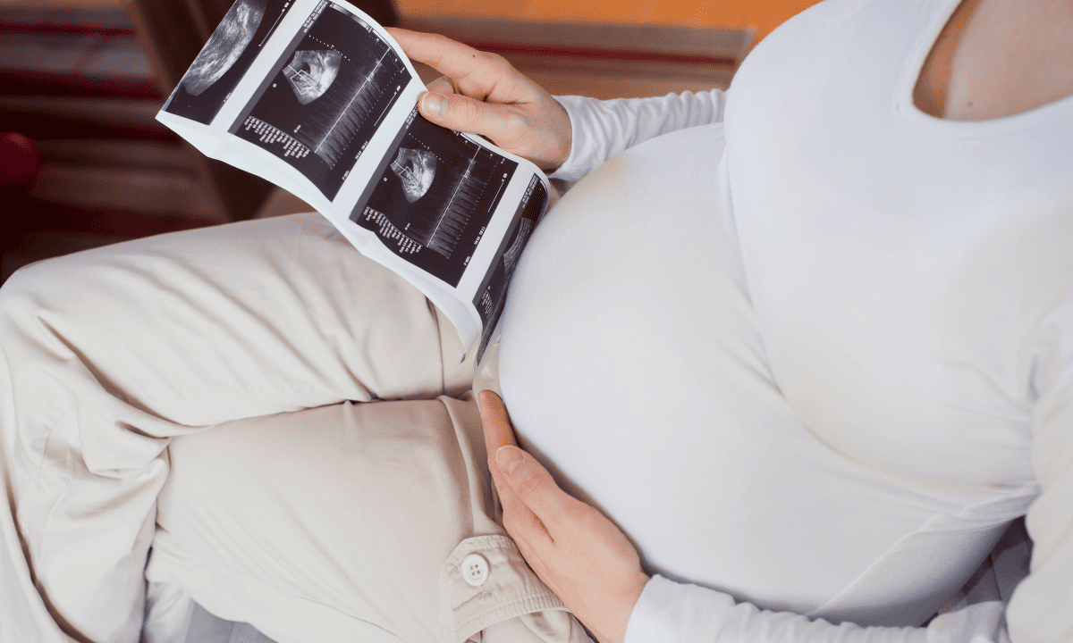 A pregnant person in a white shirt sitting down and looking at a strip of ultrasound scan photos