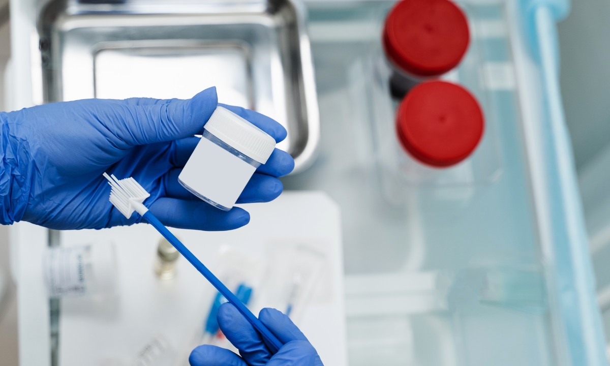 A person wearing blue medical gloves holds a small white collection container and a cervical brush/spatula, commonly used for Pap smears or HPV testing. Medical instruments are visible in the background.