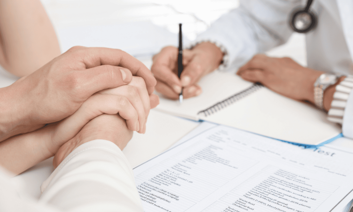 A doctor writes notes during a consultation while a patient's hands are clasped reassuringly in the foreground, signifying support during a medical discussion