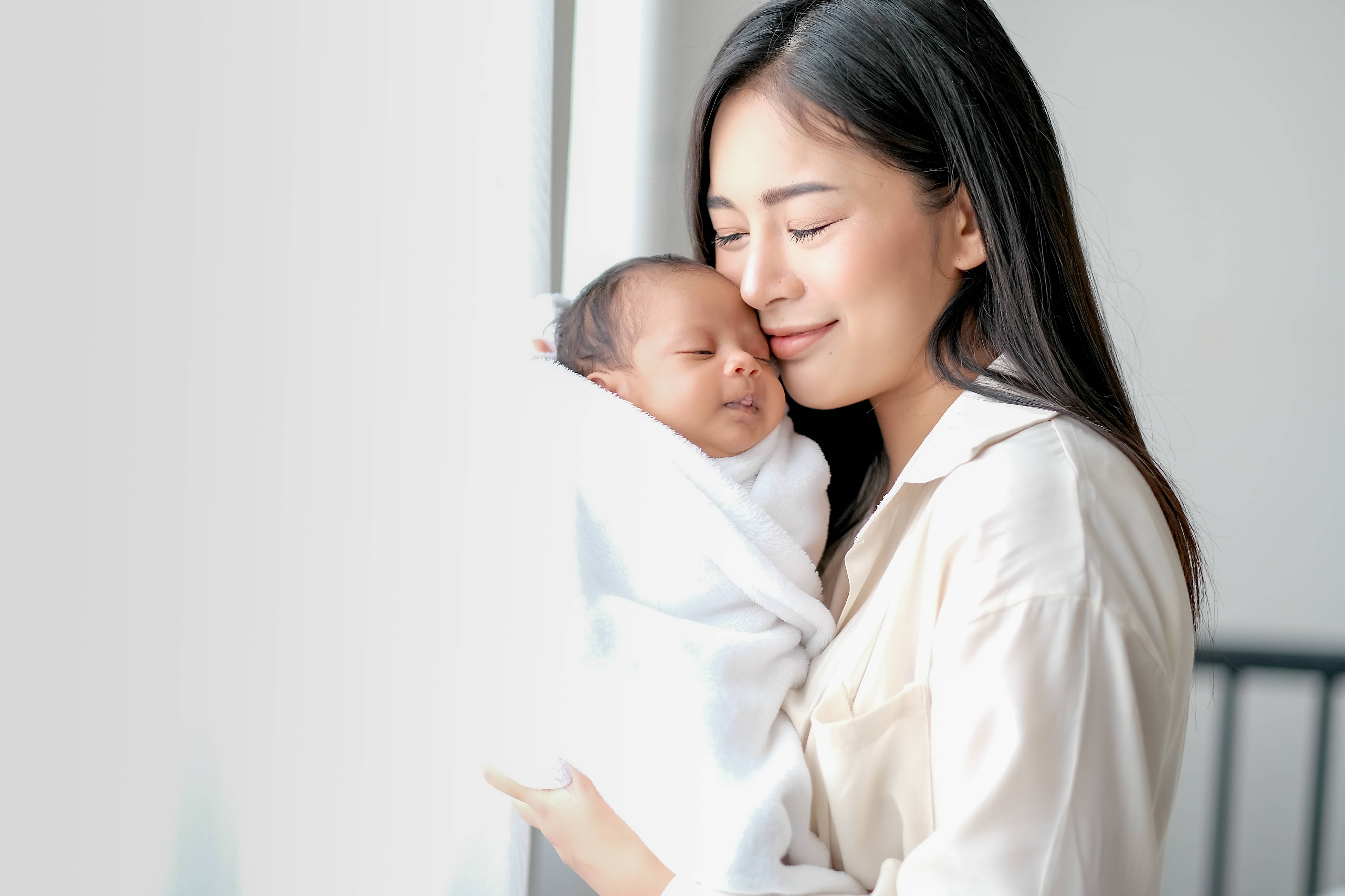 Young Asian mother cradling newborn baby while standing by the window