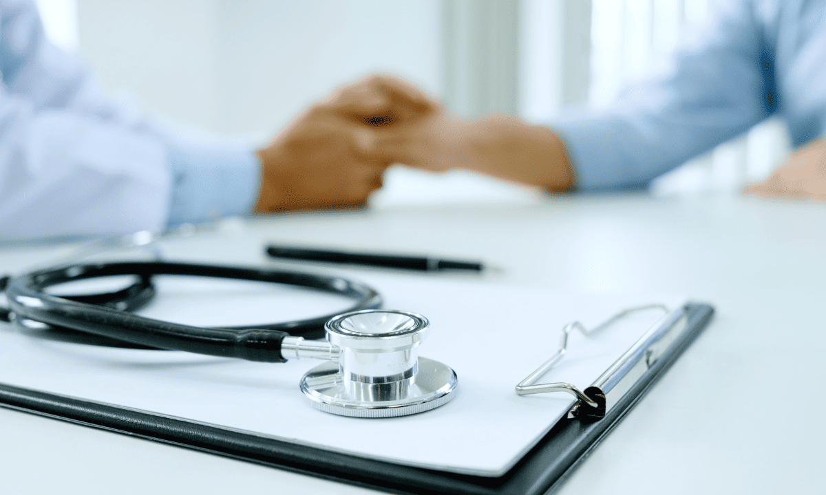 A close-up, slightly blurred photo of a doctor in a white coat holding a patient’s hand across a desk. A stethoscope and clipboard are in the foreground.