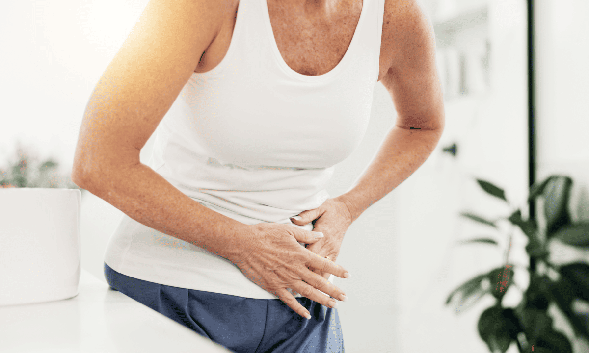 A person in a white tank top holding their abdomen in discomfort, suggesting pelvic pain or cramping.
