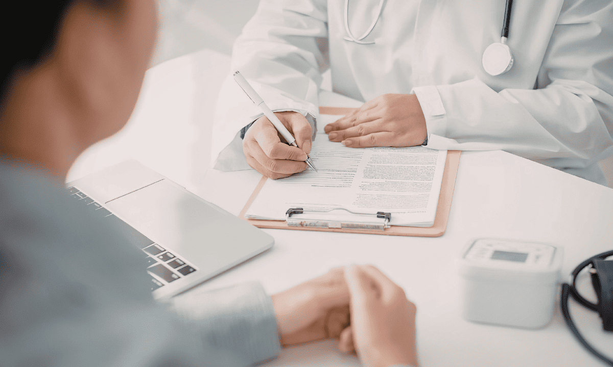 A patient sitting across from a doctor in a white coat who is writing on a clipboard during a medical consultation.