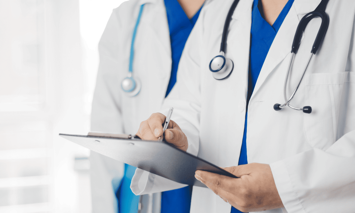 A stock photo of two doctors in white coats and blue scrubs, with one doctor holding a clipboard and pen.