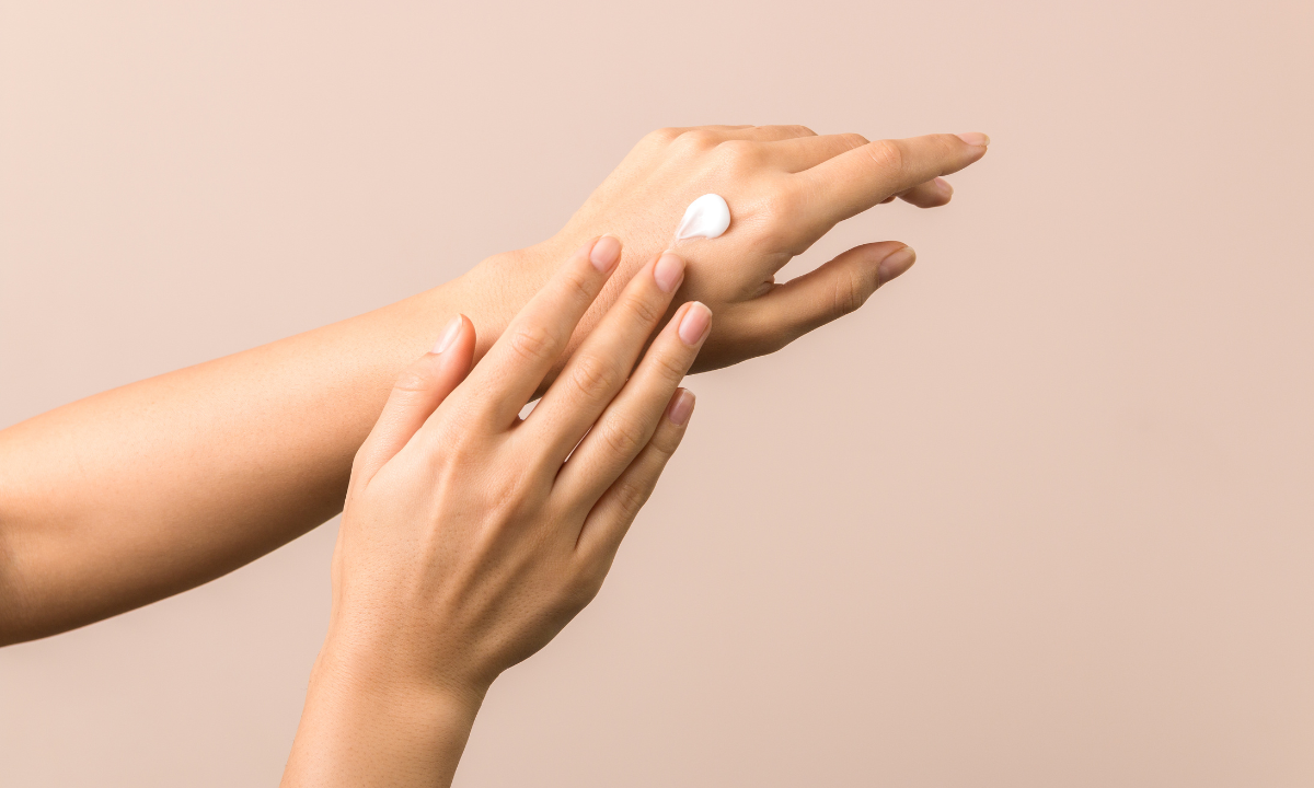 Close-up of a person's hands applying a dab of white moisturizing cream to the back of their hand.