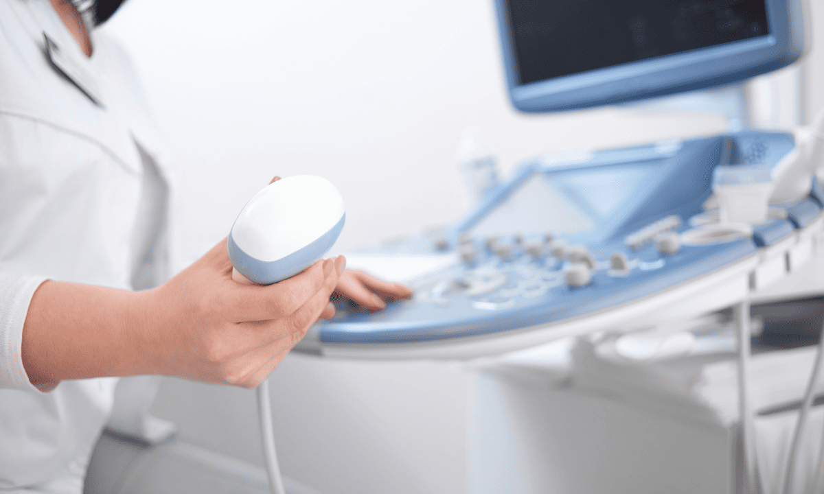A medical professional in a white coat holding an ultrasound transducer wand next to an ultrasound machine.