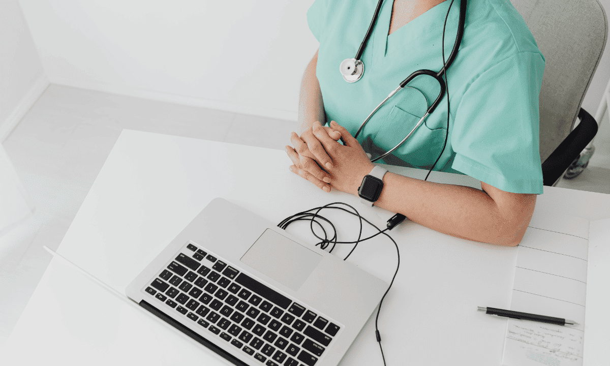 An overhead view of a healthcare professional in green scrubs sitting at a white desk with a laptop, a stethoscope, and a pen and paper, ready for a patient discussion.