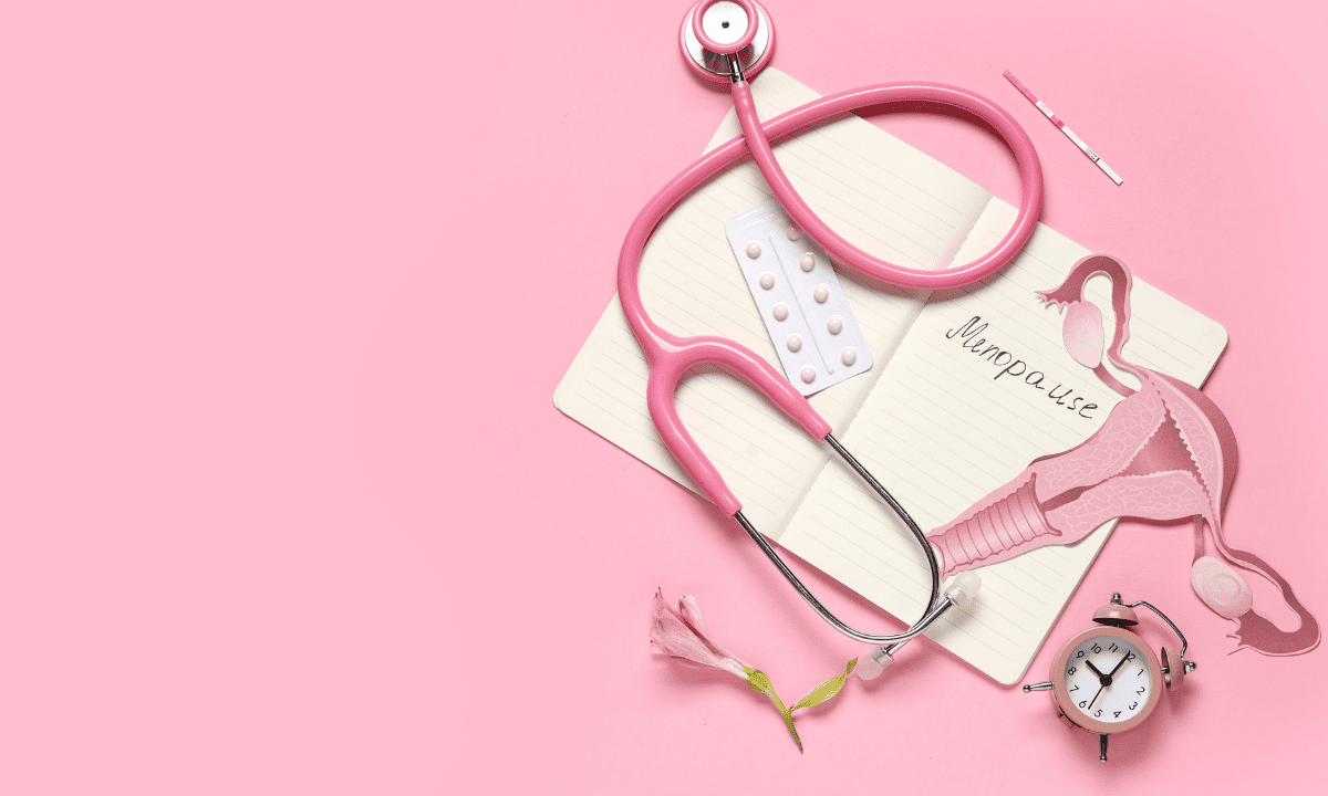 A flat-lay composition on a pink background featuring a pink stethoscope, a notebook with Menopause written in it, a blister pack of pills, and an alarm clock.