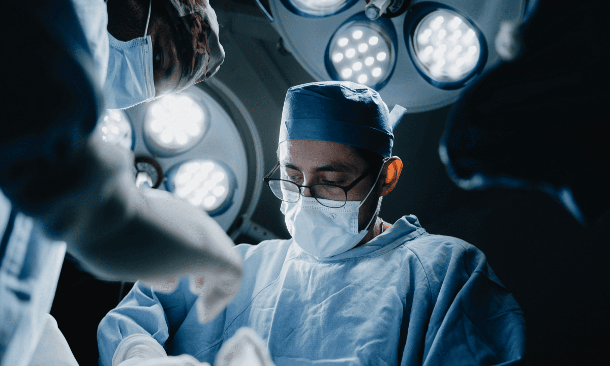 A surgeon in blue scrubs, mask, and glasses focuses intently on a procedure under bright surgical lights in an operating room.