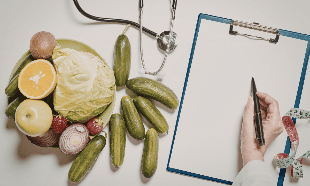 A top-down view of a doctor's hand writing on a clipboard next to a plate of fresh fruits and vegetables, a stethoscope, and a measuring tape
