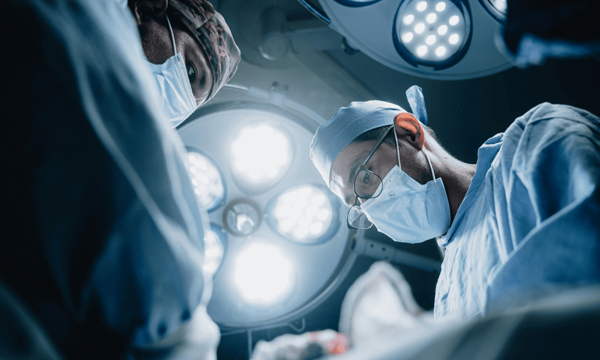 A low-angle, dramatic photograph of surgeons in blue scrubs and masks working under bright surgical lights in an operating room.