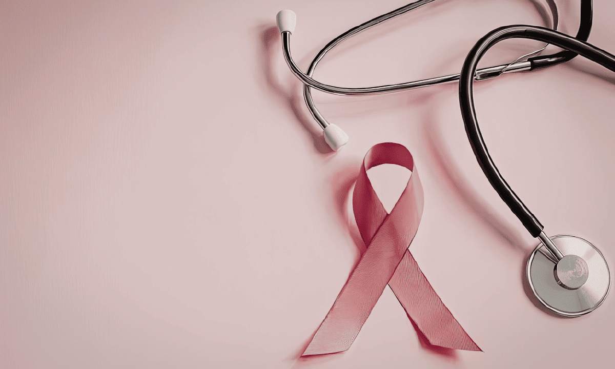 A pink breast cancer awareness ribbon lying next to a medical stethoscope on a pink background.