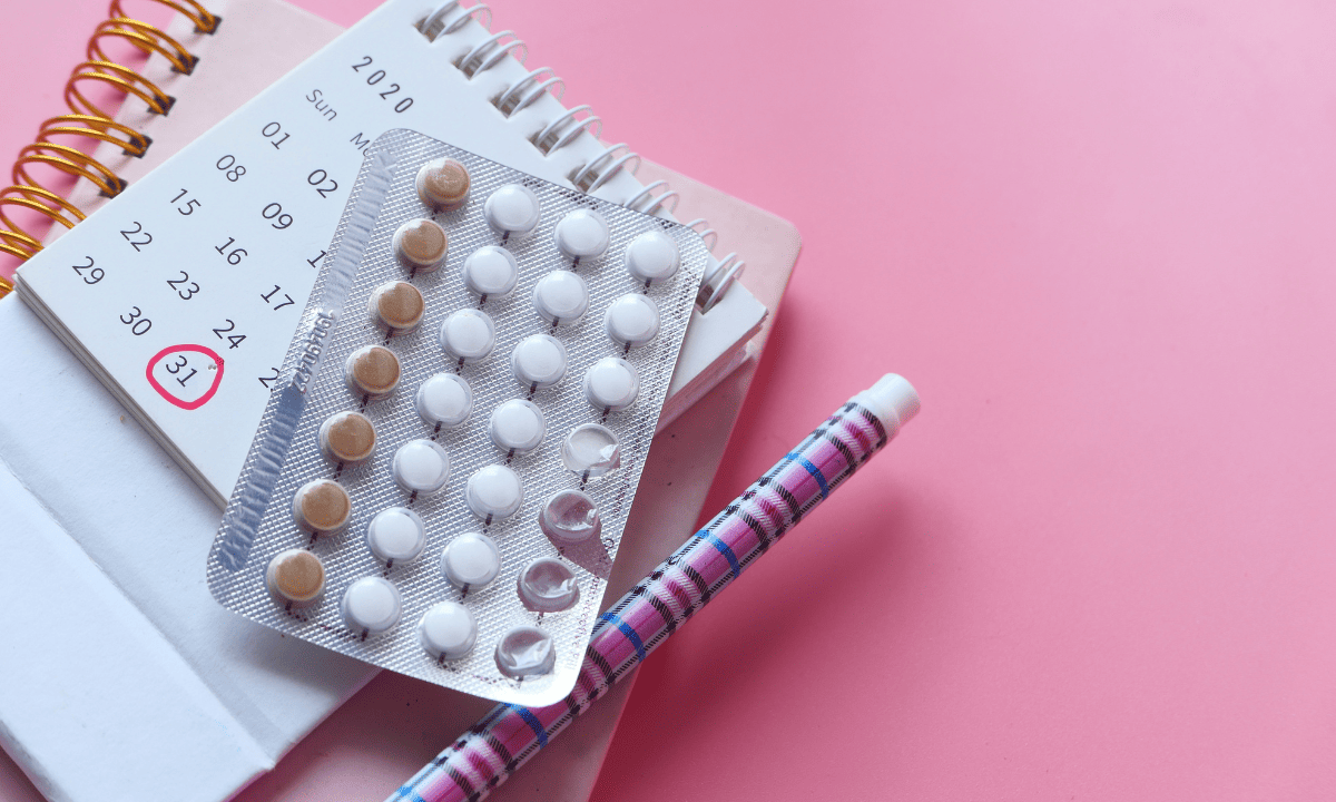 A blister pack of birth control pills resting on a small calendar and a notebook against a pink background, symbolizing hormonal treatment or cycle tracking.