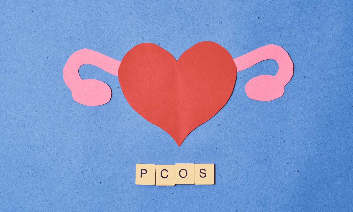 A paper craft illustration showing a red heart as the uterus with pink fallopian tubes, accompanied by letter tiles spelling PCOS on a blue background.