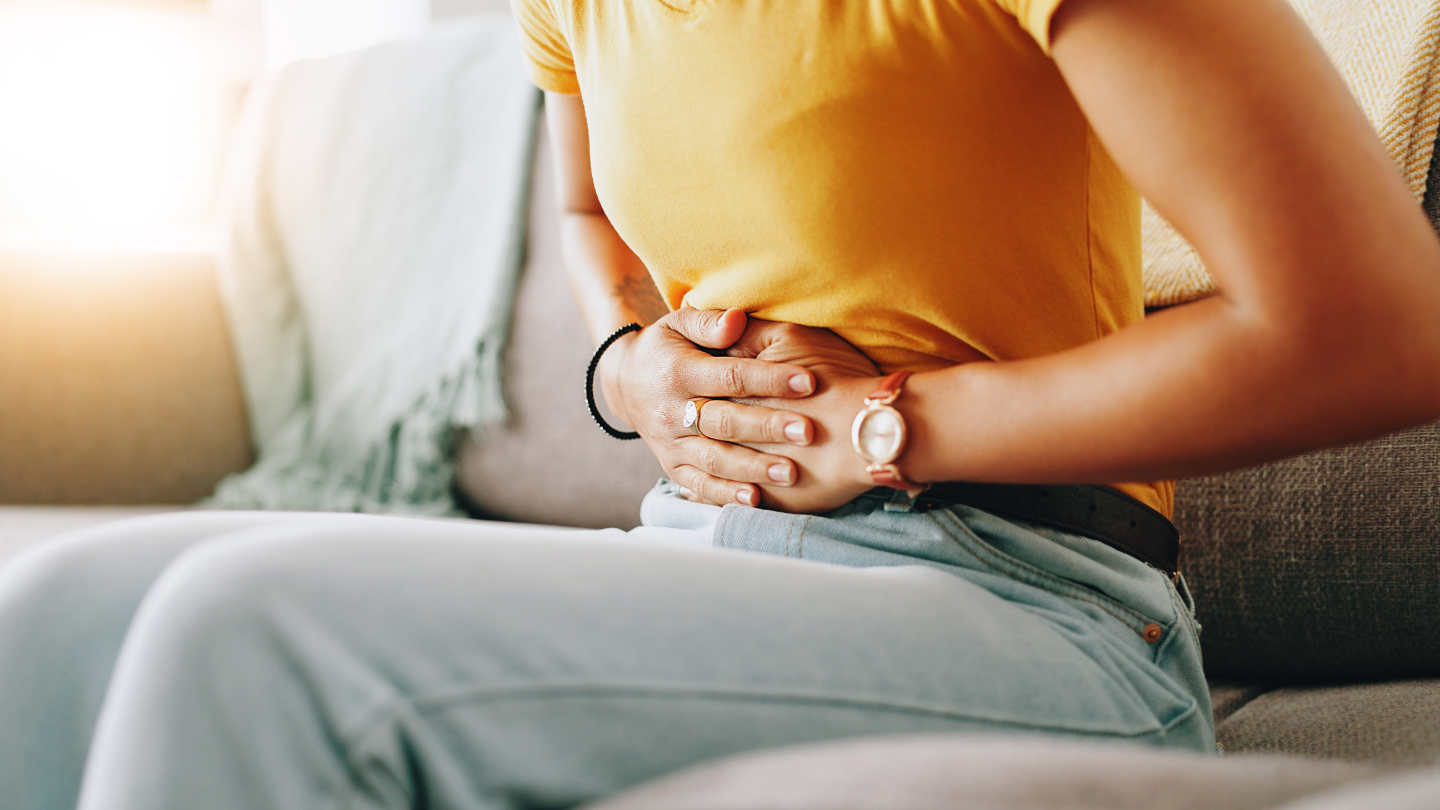 Person seated on a sofa clutching their lower abdomen, showing abdominal discomfort.