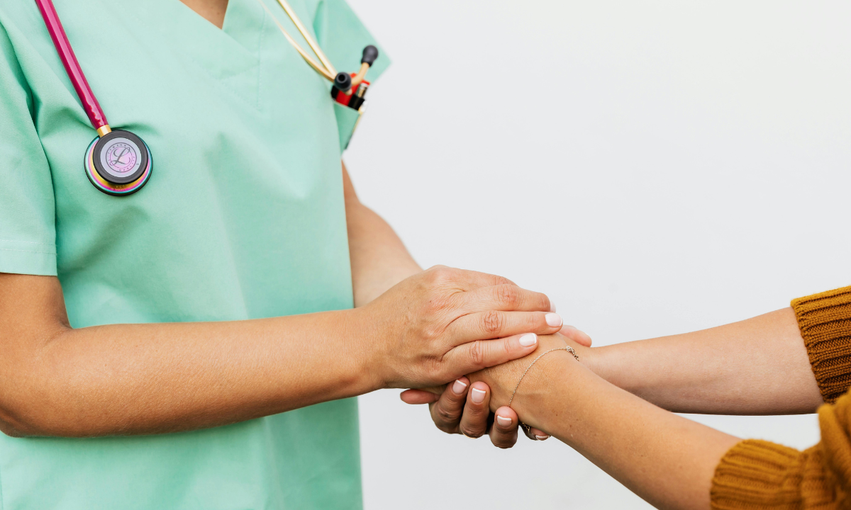 Close-up of a healthcare provider in light green scrubs holding a patient's hand with both hands, providing comfort and reassurance. The provider is wearing a stethoscope.