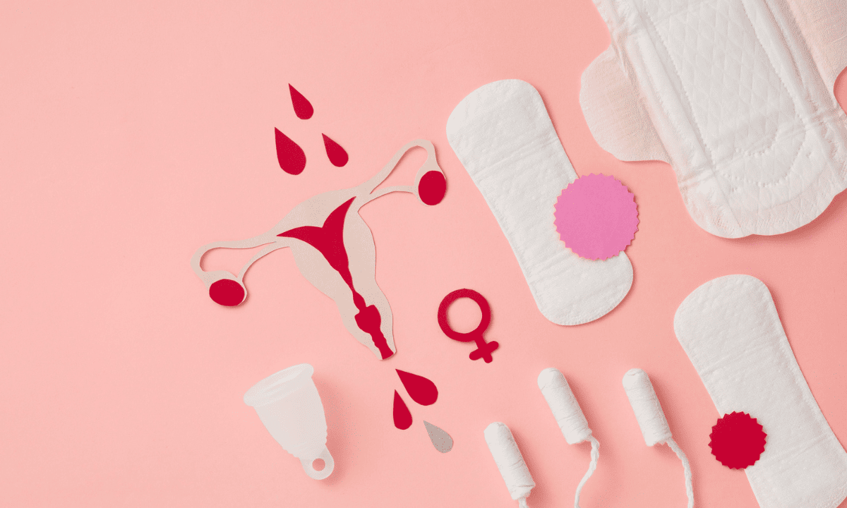 An overhead flat lay of various menstrual products arranged on a pink background, including pads, tampons, a menstrual cup, and a paper cutout of a uterus