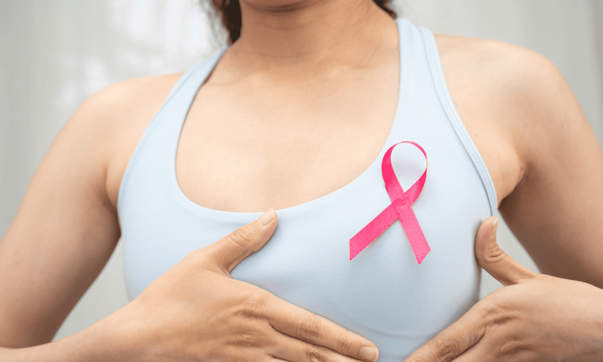 A woman in a sports bra holding her hand near a pink breast cancer awareness ribbon pinned to her chest.