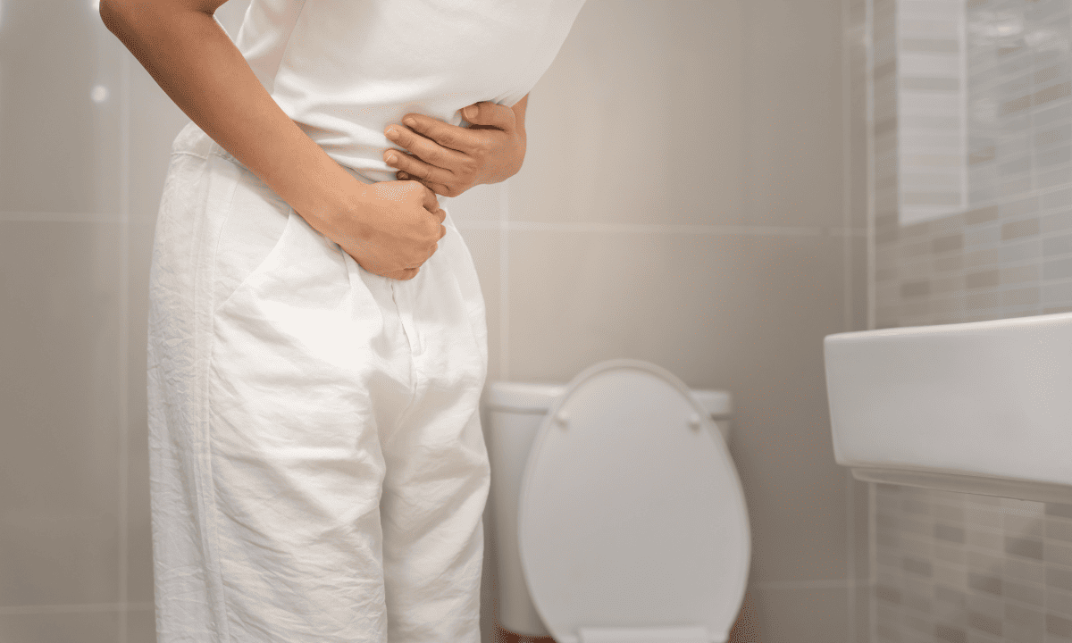 Person in white clothing standing in a bathroom, clutching their stomach in pain.