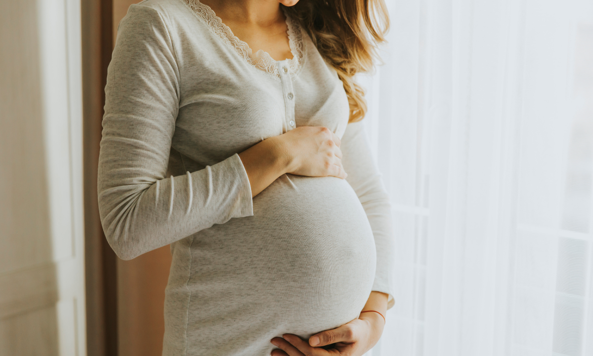 A pregnant woman in a white dress standing by a window, gently holding her baby bump.