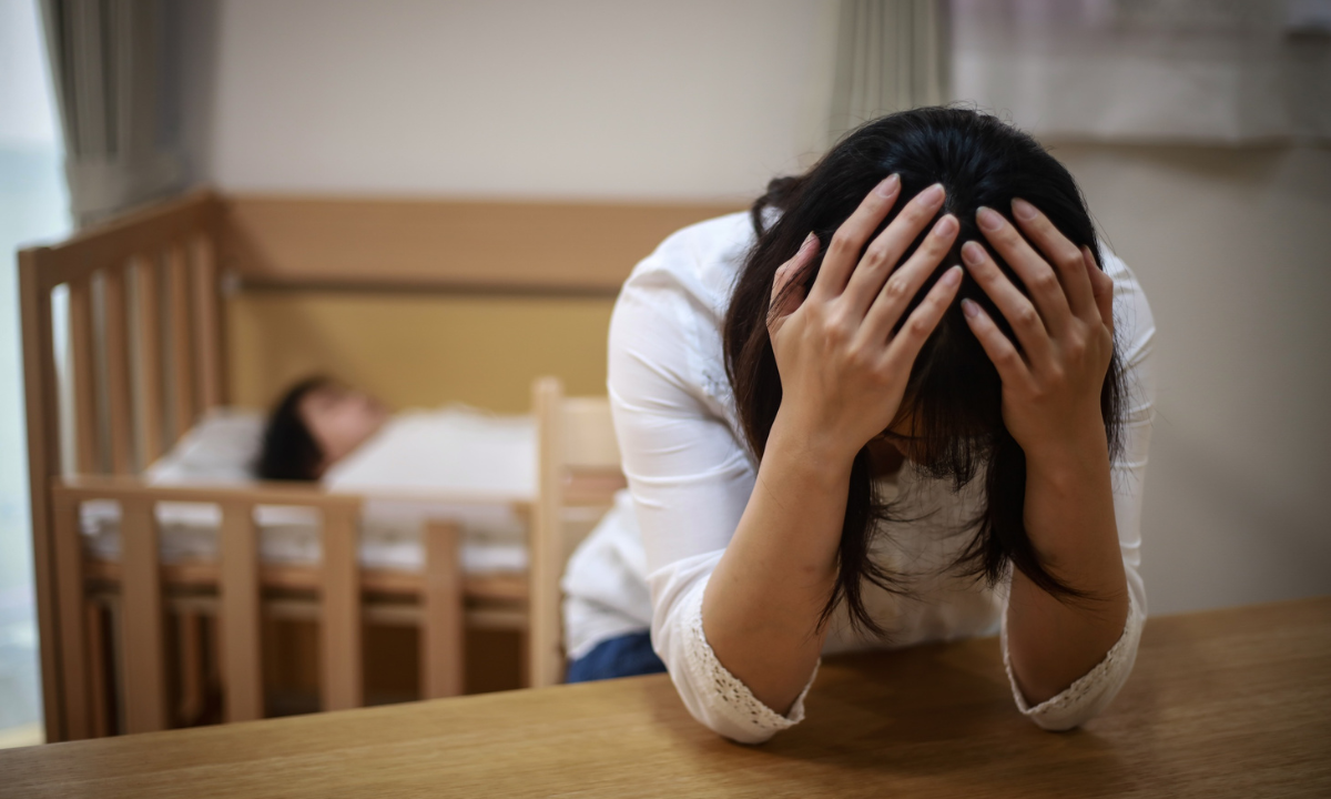 A stressed mother sits with her head in her hands while her baby sleeps in a crib in the background.