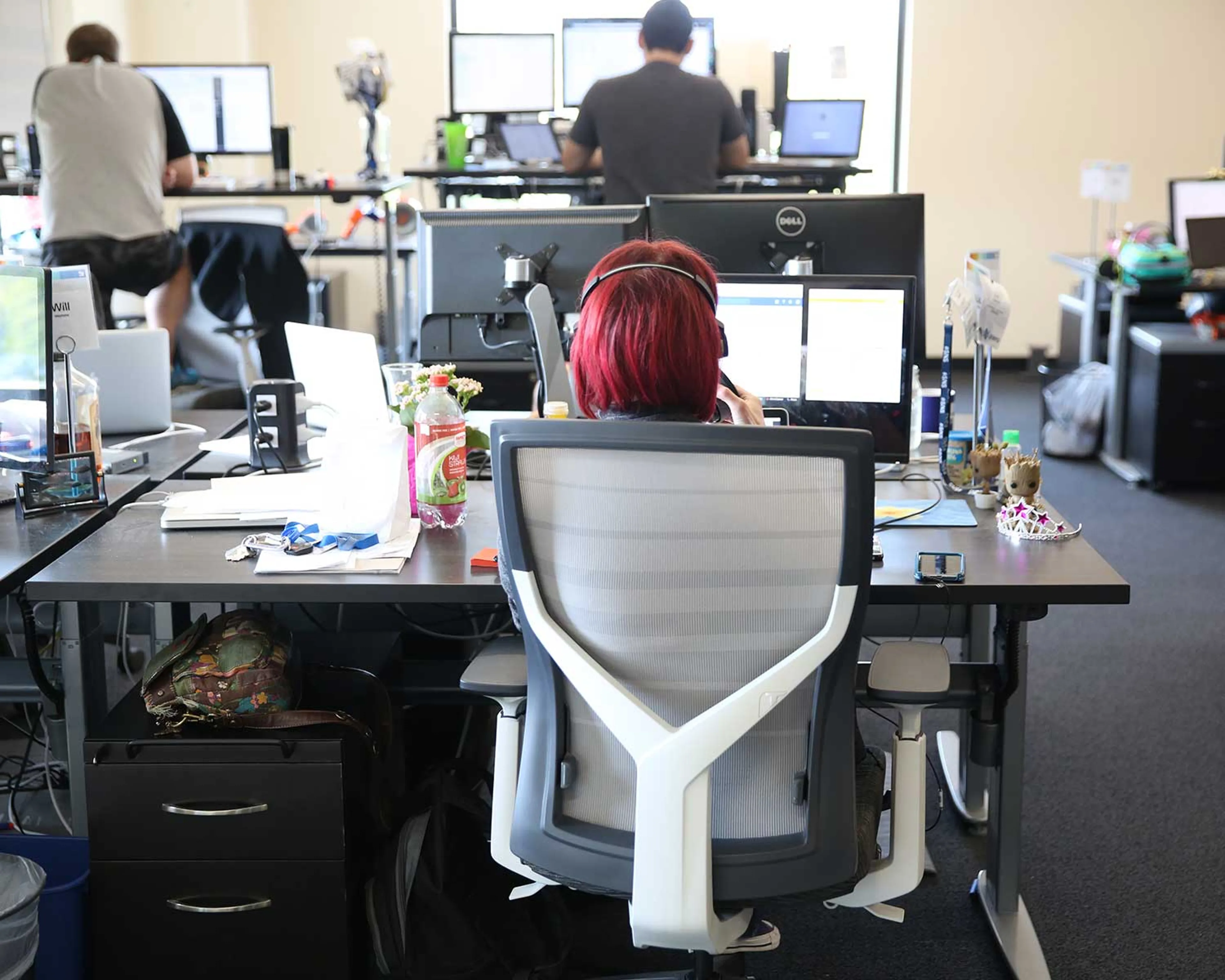 A person with short, bright red hair is sitting in an office chair at a desk with multiple computer monitors, working in a busy office environment.