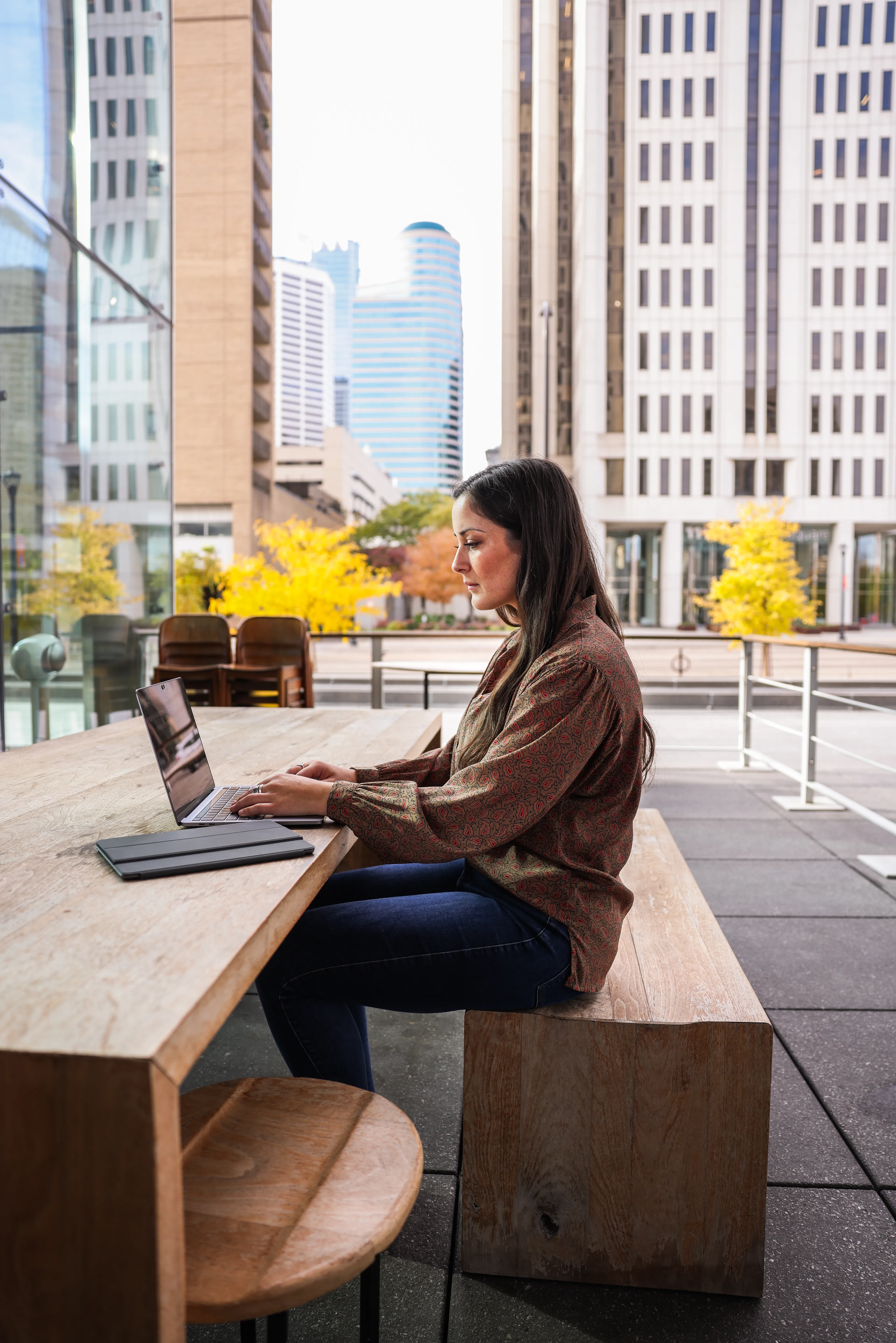 A woman with long dark hair is working on a laptop at a wooden table outdoors in a city setting.