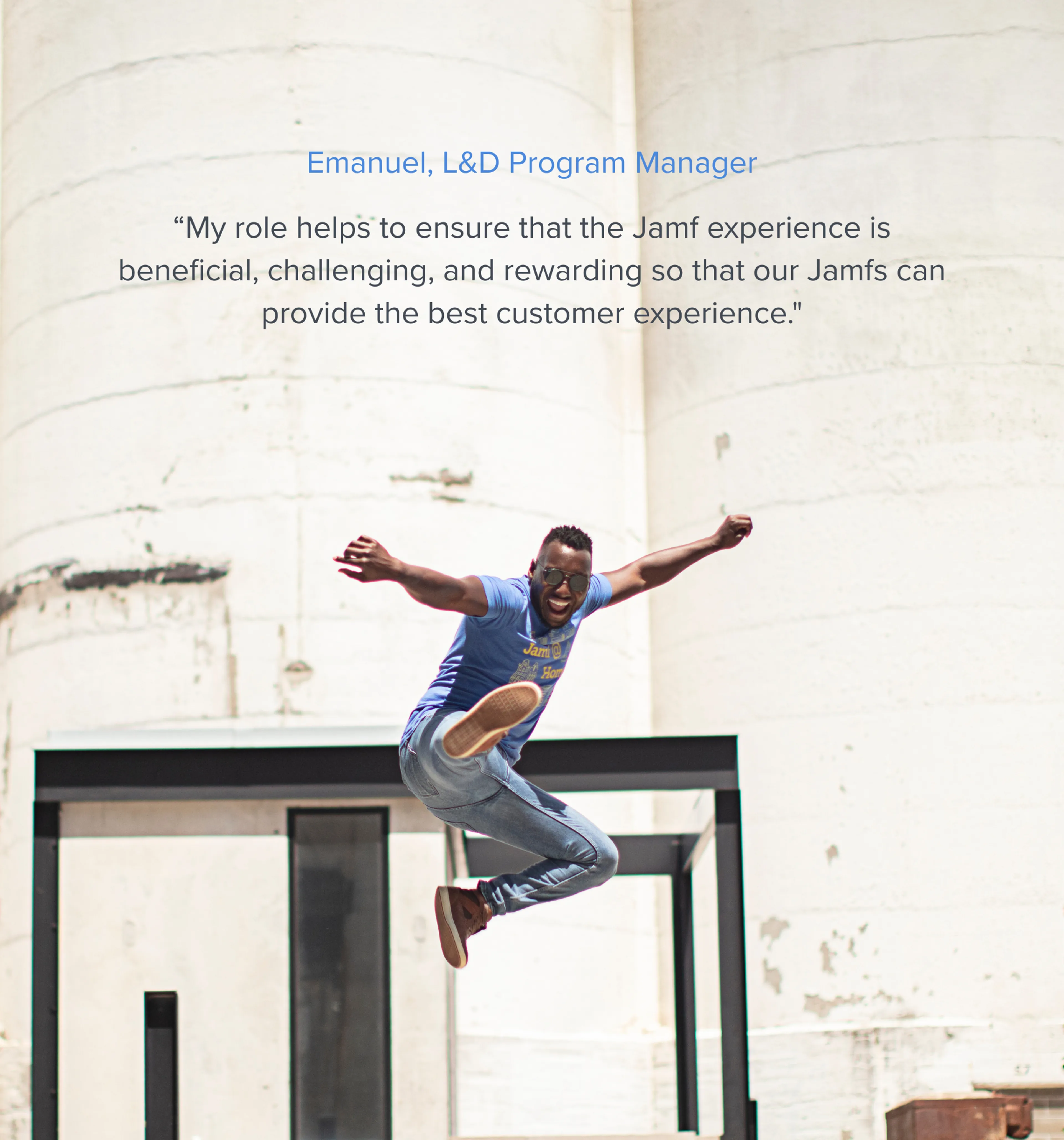 A smiling Learning and Developement Program Manager, leaps joyfully in a blue "Jamf" t-shirt, jeans, and sunglasses against a backdrop of large concrete silos and a dark architectural structure.
