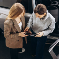 Two people standing beside an open car door, looking at and discussing a tablet device.
