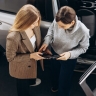 Two people standing beside an open car door, looking at and discussing a tablet device.
