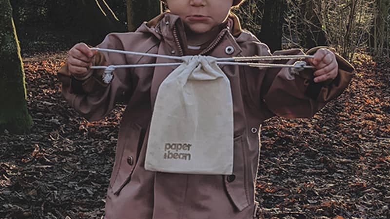 Toddler dressed in woolly hat and boots in the woodland between trees with sun rays shining through. Toddler is holding a Paper & Bean cotton bag.