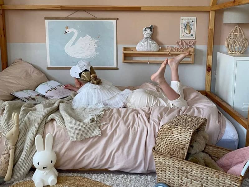 Looking in on a girl laying on her house frame bed reading. On the floor is a Miffy lamp and an Olli Ella Strolley. The wall is decorated with a wide horizontal