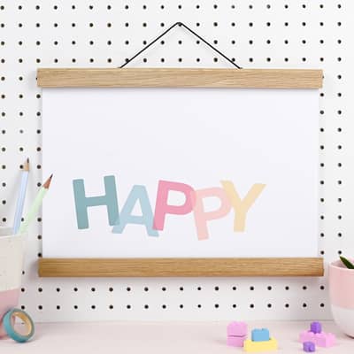 An art print hangs in a wooden poster hanger. The background is white and there are pastel letters spelling out the word HAPPY.