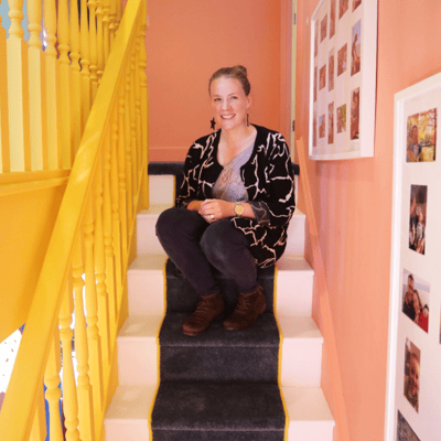 A white woman with brown hair sits on her stairs. The walls are coral pink and the bannisters are painted bright yellow.