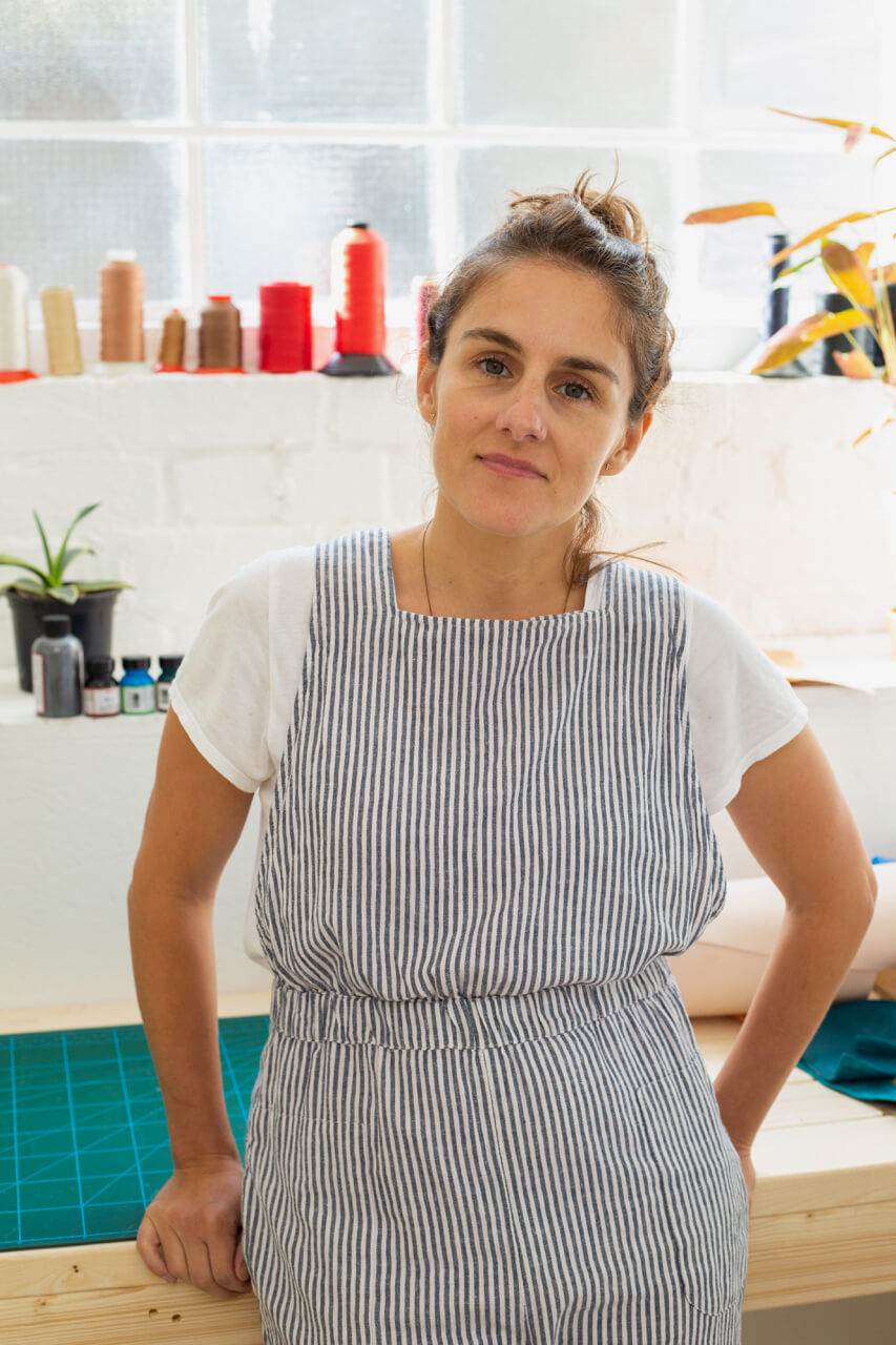 White woman smiling and leaning against workbench in sewing studio