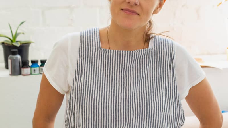 White woman smiling and leaning against workbench in sewing studio